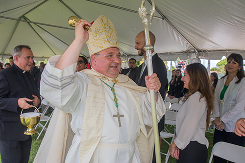 Auxiliary Bishop Peter Baldacchino sprinkles holy water on St. Brendan staff and students during the groundbreaking for the high school's new Innovation Center. At left is Father Jose Alvarez, president of St. Brendan High and pastor of Our Lady of the Lakes Parish in Miami Lakes.