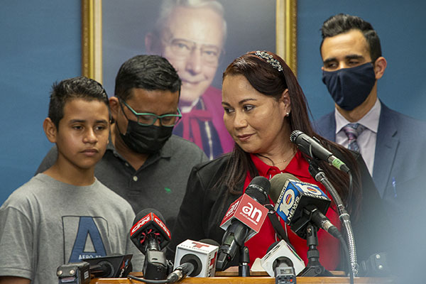 Durante una conferencia de prensa el 10 de febrero de 2022, el abogado de Catholic Legal Services, Alberto Montequin, al fondo a la derecha, observa cómo Yesenia, que vive en Miami, expresa su gratitud a Msgr. Bryan O. Walsh Children's Village, de Caridades Católicas, donde su hijo, Andy, al fondo a la izquierda, pasó 11 días antes de reunirse con ella tras cruzar la frontera desde Honduras. También mira su padrastro, Manuel. Los refugios de toda La Florida como el que dirige Caridades Católicas, corren el riesgo de tener que cerrar debido a una orden ejecutiva del gobernador Ron DeSantis.