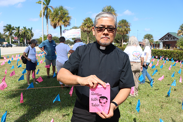 El P. Edgardo De Los Santos sostiene una banderita en memoria de un bebé que murió durante un aborto. Un total de 3,000 banderas azules y rosas se desplegaron en los terrenos de la parroquia St. Ambrose, en Deerfield Beach, durante todo el mes de octubre, como parte de un proyecto del Mes del Respeto a la Vida del Ministerio BLESS por la Vida de la parroquia.