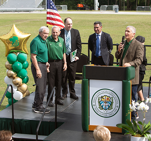 Audience applauds Coaches Van Parsons and Carmen Grosso, standing on stage, for whom the new athletic facility is named, as Jim Rigg, Miami Mayor Francis Suarez and Ignacio Halley, class of '78, look on, during the dedication and blessing of new athletic facilities at Immaculata LaSalle High, Feb. 1, 2024.
