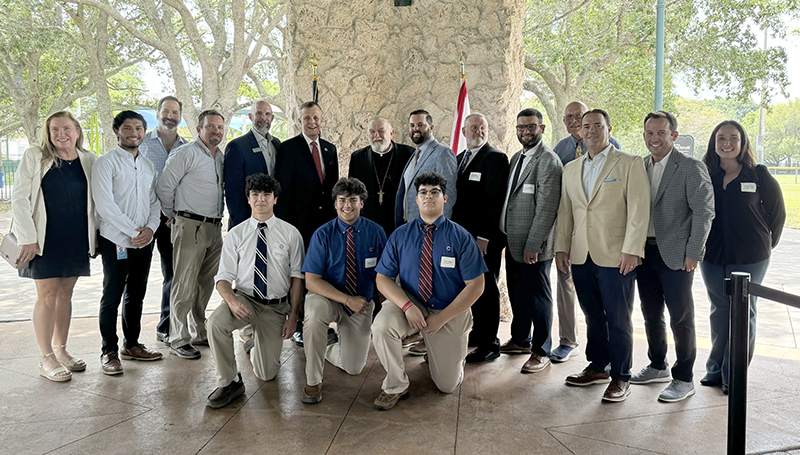 Archbishop Thomas Wenski (center) poses with Thomas G. Kruczek, president of Columbus High School (directly left of archbishop); Nick Ramos, Columbus vice president of advancement (directly right of archbishop); and a group of Columbus staff, students, and alumni May 15, 2024, before the start of the Miami’s Community News Pinecrest Luncheon with Archbishop Thomas Wenski” at Evelyn Greer Park, in Pinecrest.