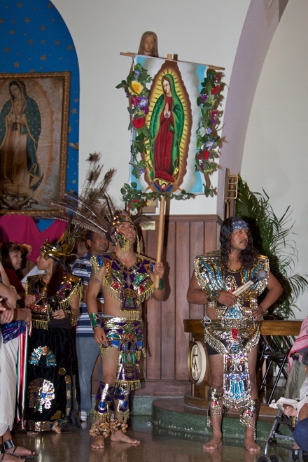 Representatives of the Aztec community process out of Little Flower Church after Mass while dancing and holding a flag with the image of Our Lady of Guadalupe.