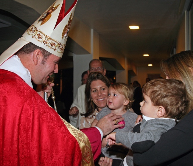 Chorbishop Michael Thomas greets Juliana Rose Romano, 17 months, as her mother, Katie Romano, holds her. Matthew Bennett, also 17 months, and his mother, Helen Bennett, look on